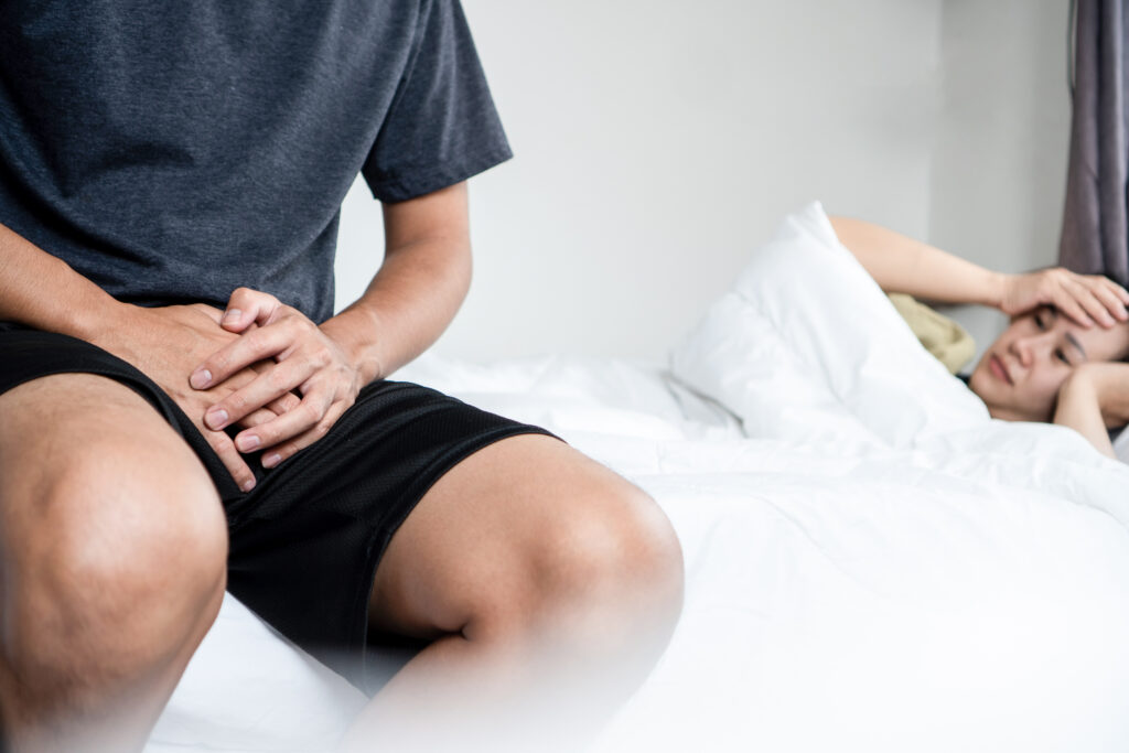 Man sitting on edge of bed with hands over groin while wife lies in bed